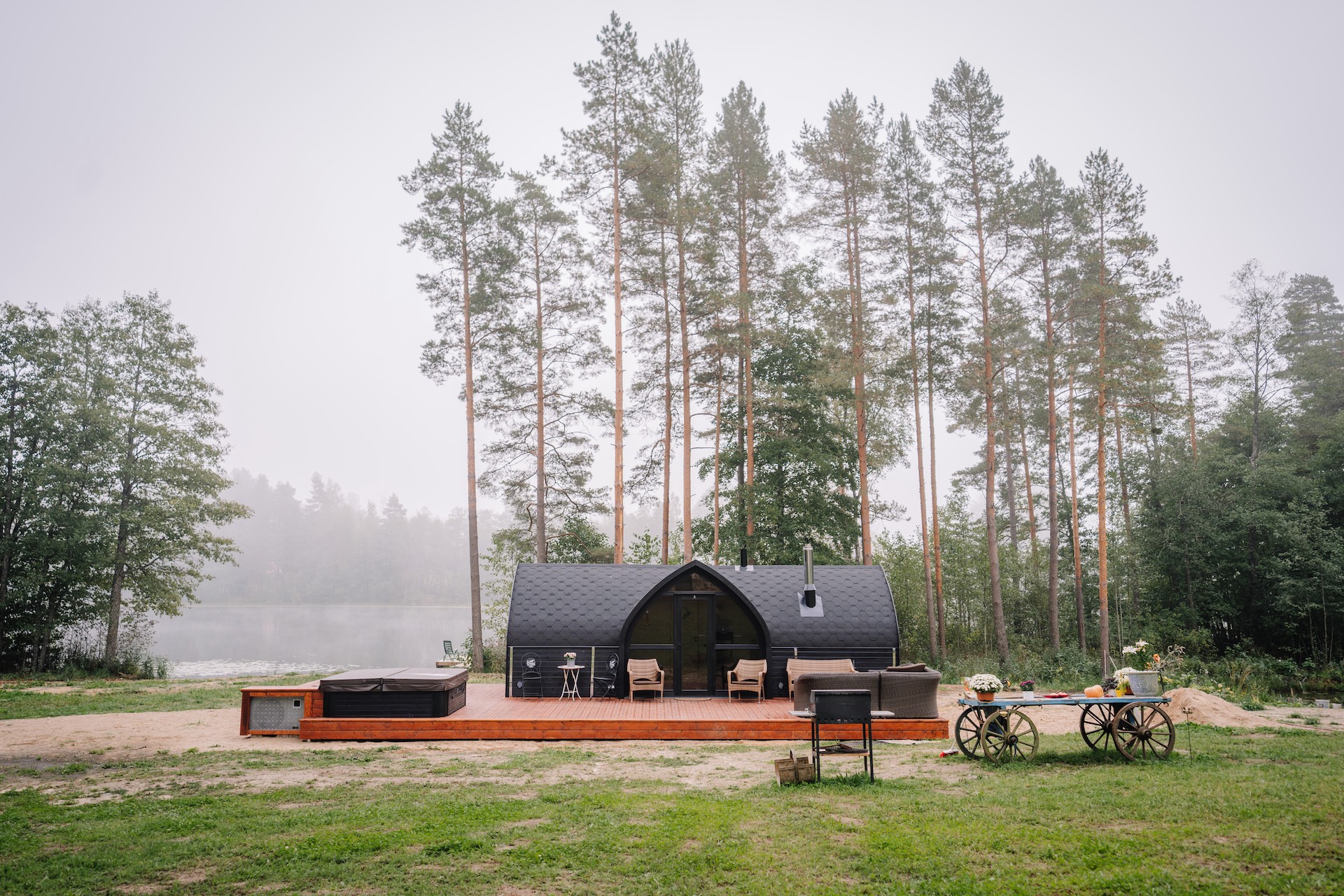 A modern cabin with a hot tub on a wooden deck, set by a lake and surrounded by tall trees, featuring outdoor furniture and a rustic table.
