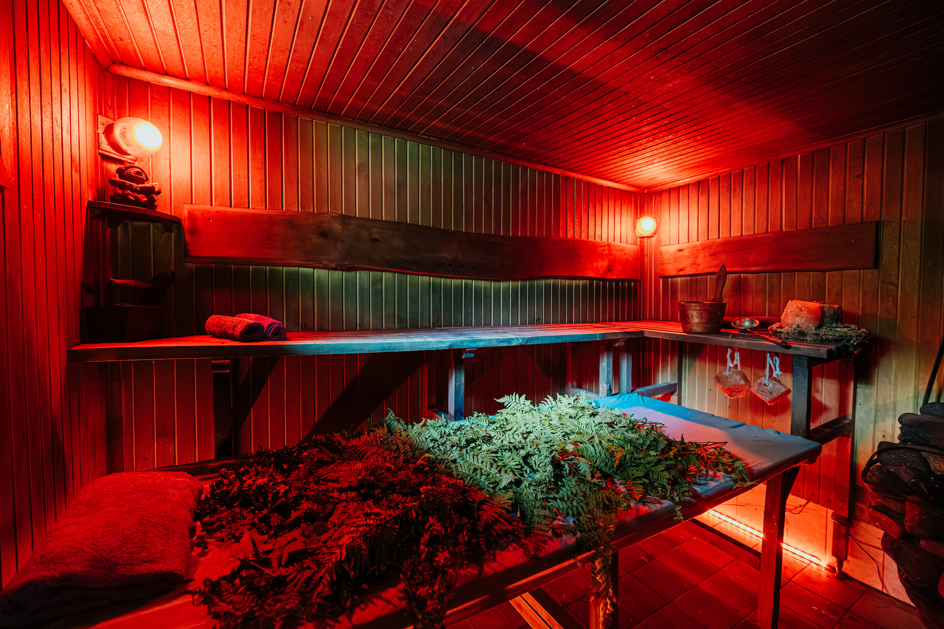 Rustic wooden sauna room bathed in red ambient light, featuring herbal venik bundles, wooden benches, towels, bucket, and wellness items for a traditional steam ritual